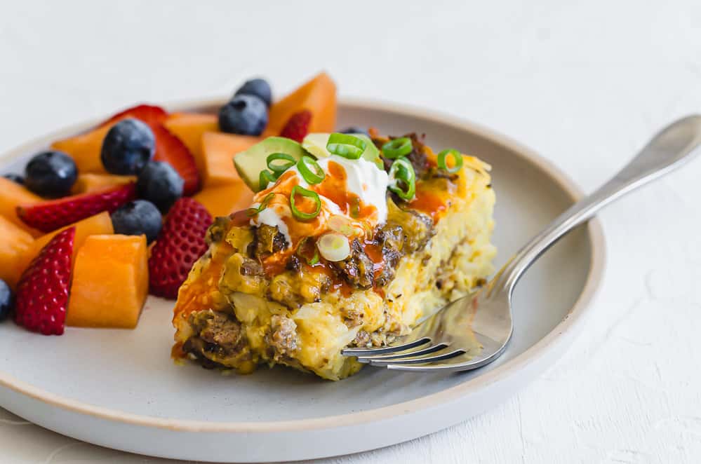 A slice of breakfast casserole with hash browns on a plate with a fruit salad