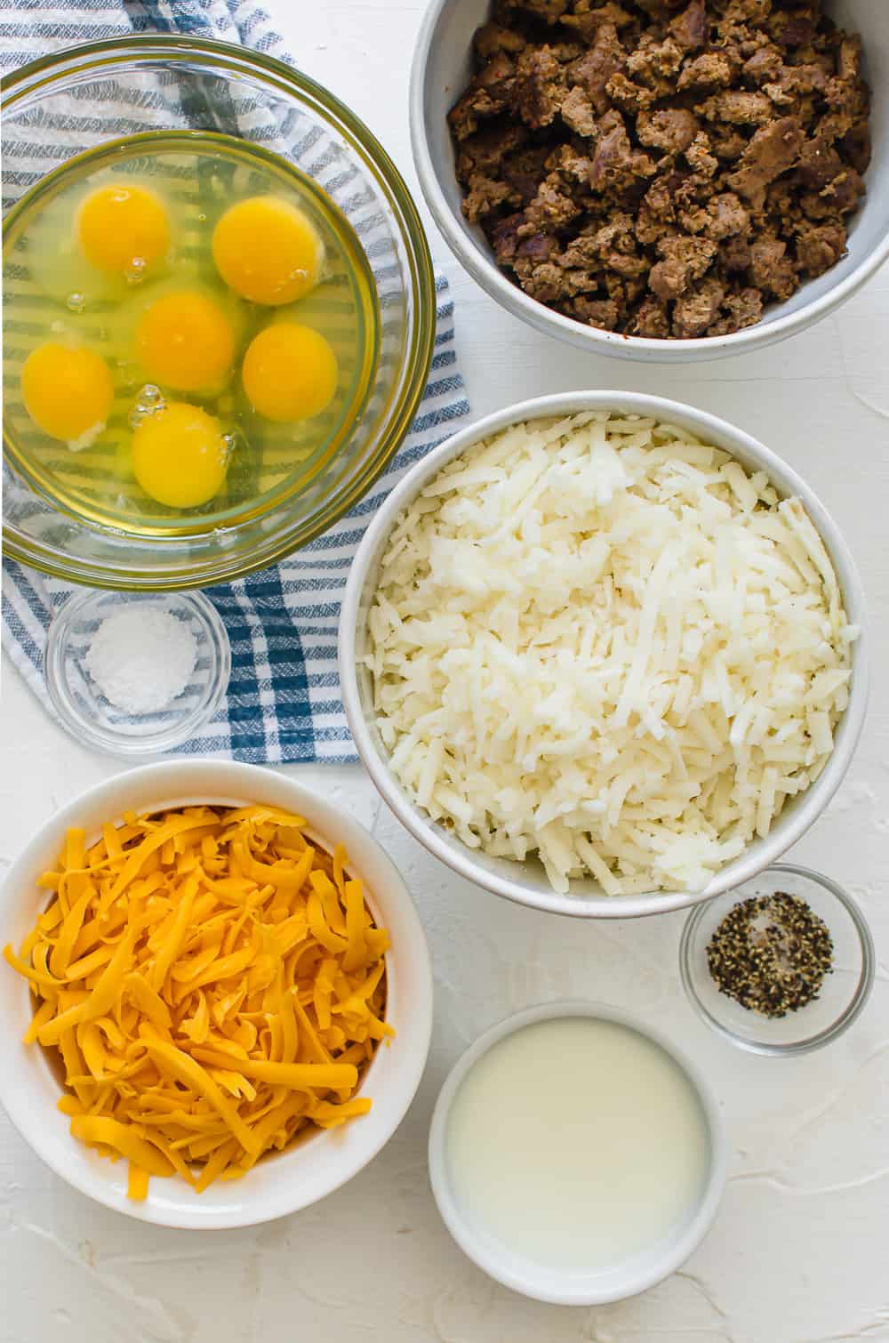 ingredients for a breakfast casserole, including eggs, browned sausage, hash browns, shredded cheese, milk, salt and pepper