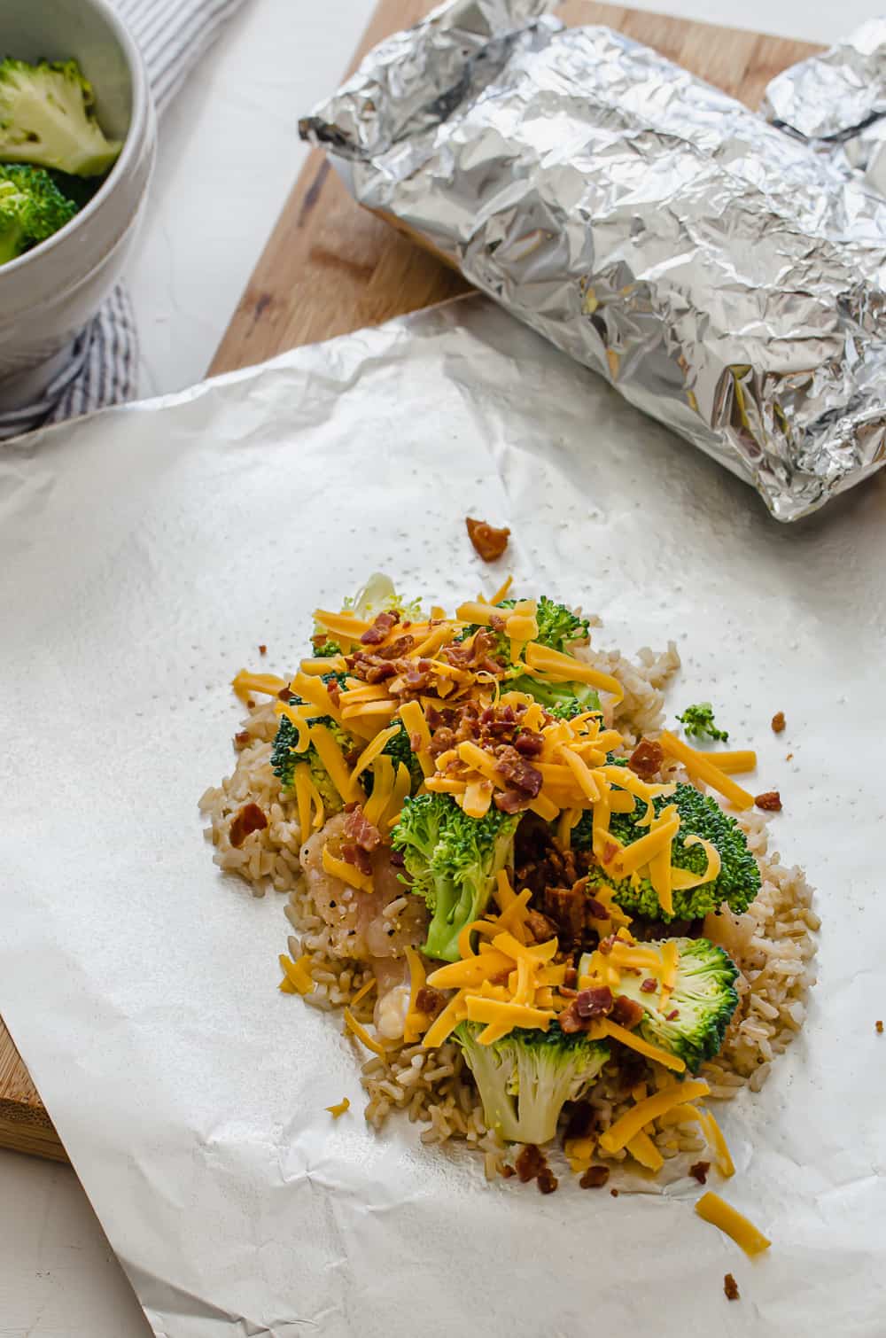 Cooked rice, raw diced chicken, broccoli, cheese, and cooked bacon on a piece of foil to be made in a packet.