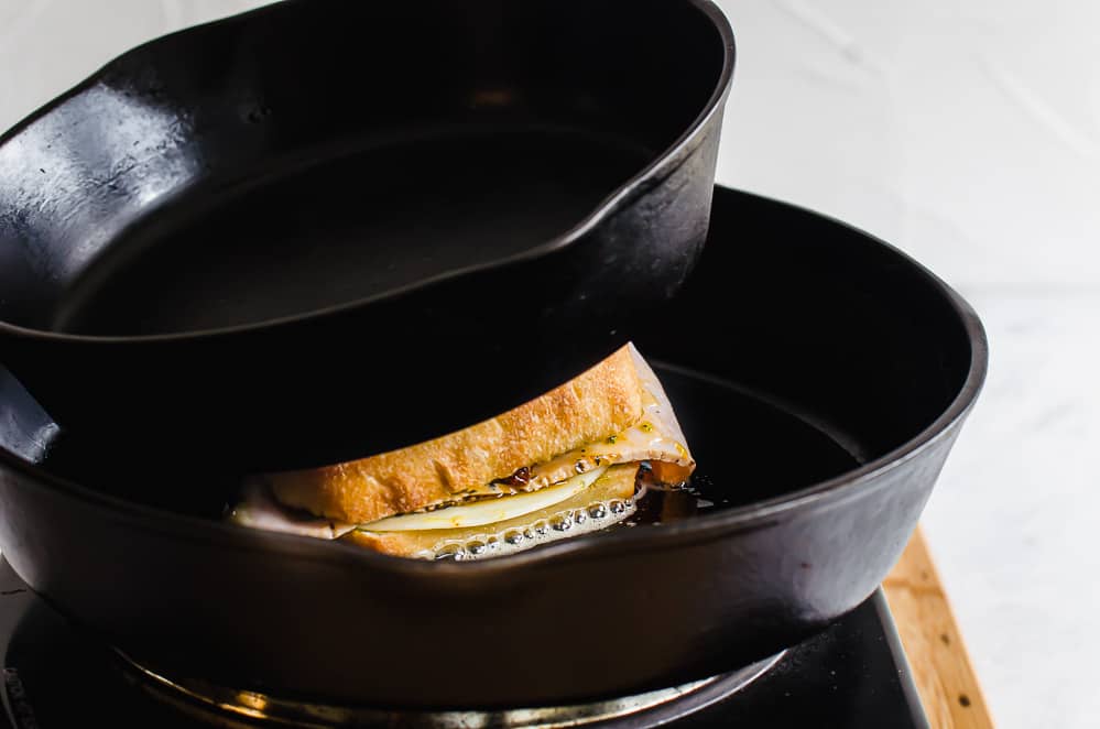 A turkey pesto panini in a cast iron skillet with another cast iron skillet on top of the sandwich pressing it down.