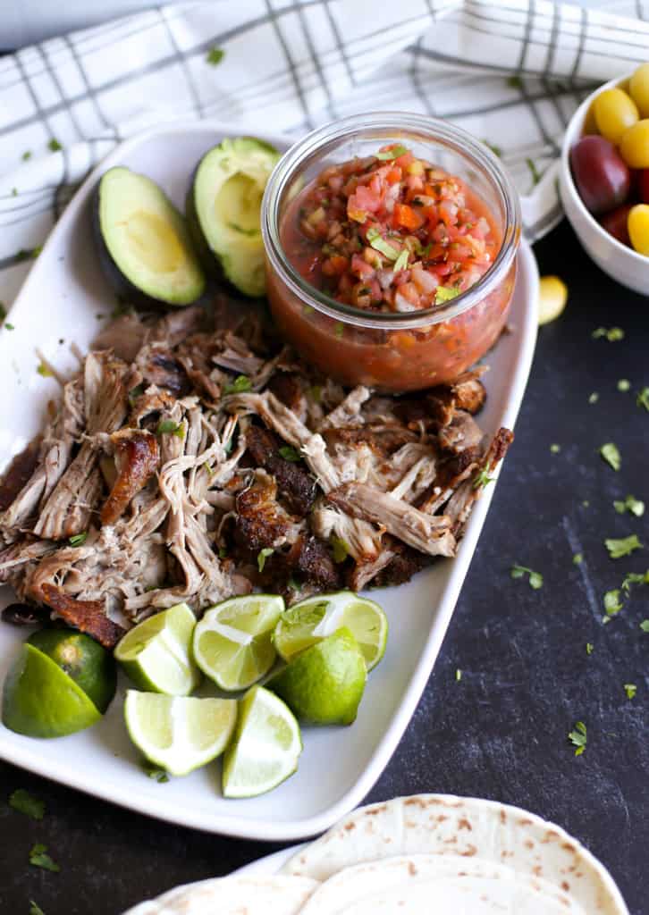 Shredded pork carnitas on a white platted with lime wedges, avocado halves, and a small jar of salsa.