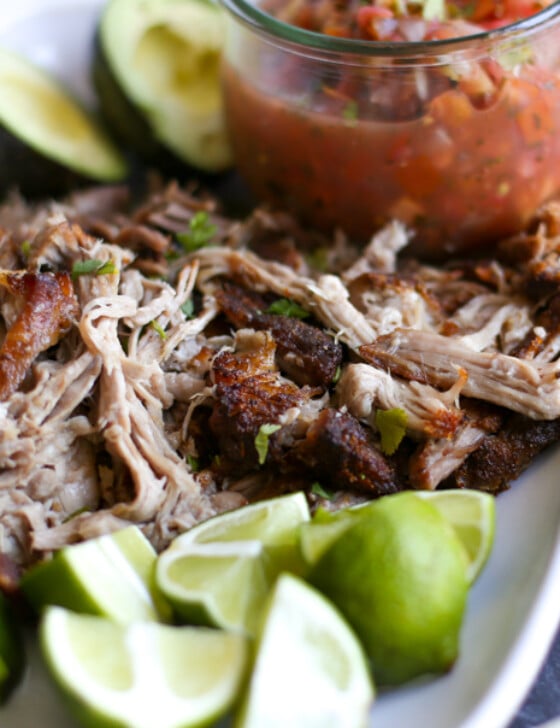 Carnitas served on a white platter with a small jar of salsa, lime wedges, and avocado halves.