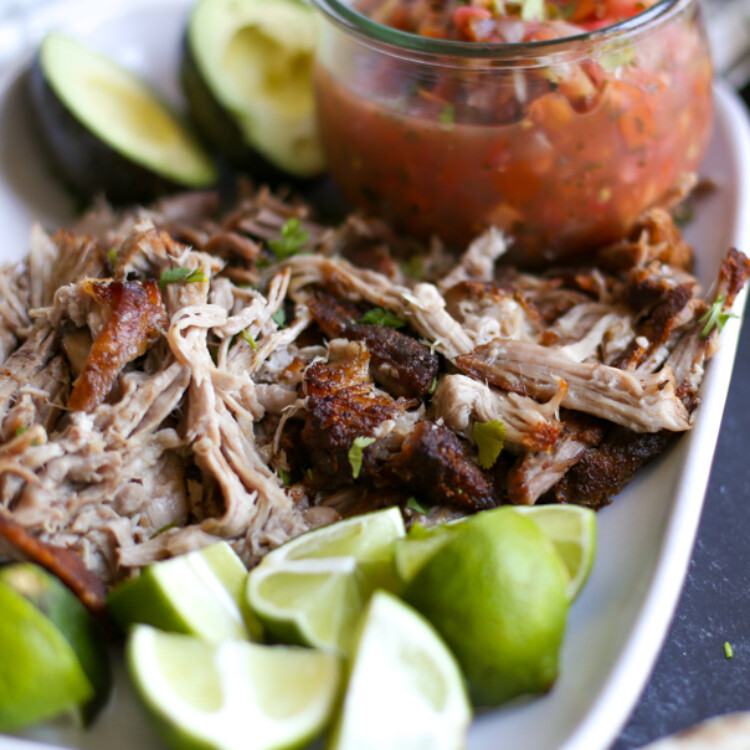 Carnitas served on a white platter with a small jar of salsa, lime wedges, and avocado halves.