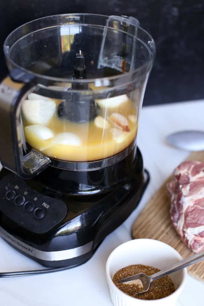 Carnitas marinade in a food processor  with a raw pork shoulder and bowl of seasonings next to it.