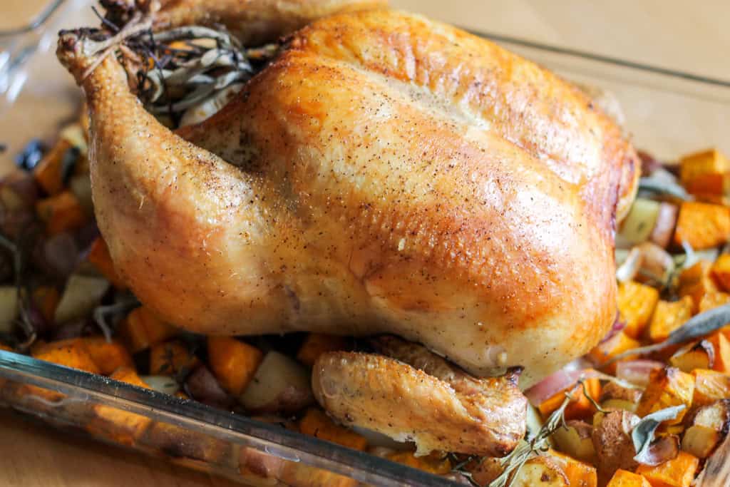 A whole roasted chicken on top of vegetables in a glass baking dish. 