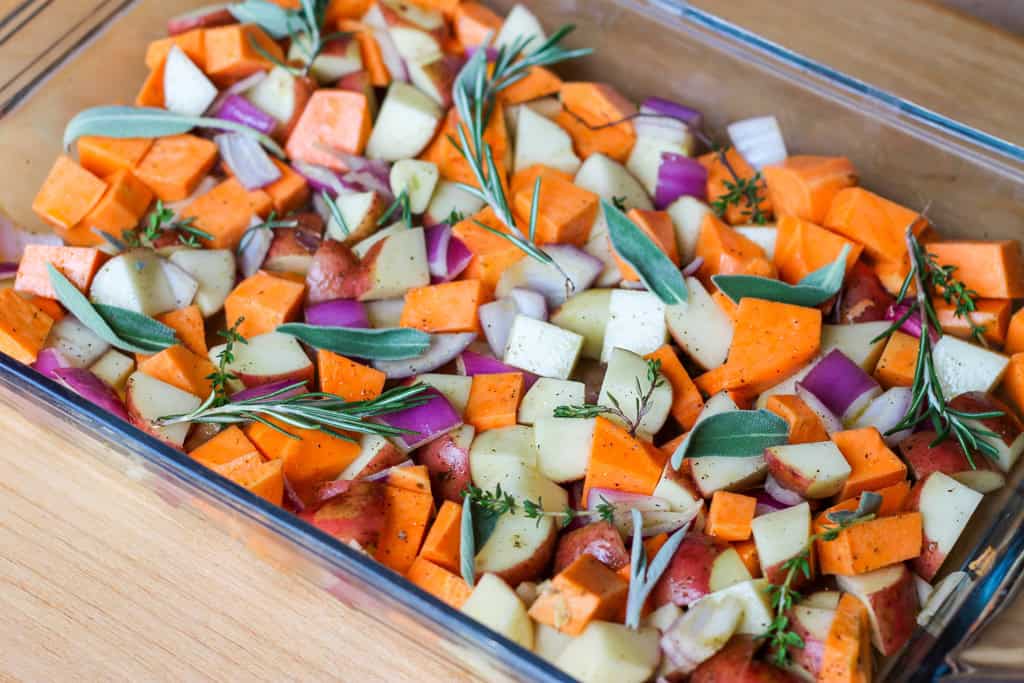 Chopped up root vegetables like sweet potato, red potato, and onion with rosemary and sage in a roasting pan.