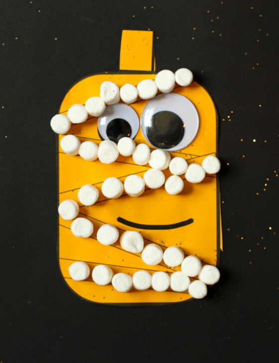 Pumpkin cut out of orange paper glued on a piece of black paper with googly eyes and mini marshmallows in lines to make it look like a mummy.