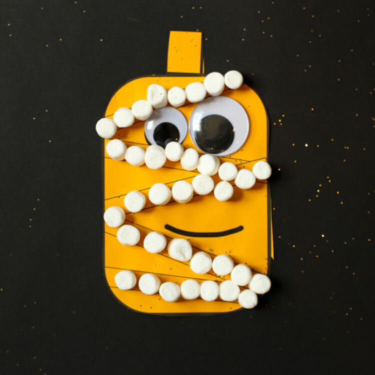 Pumpkin cut out of orange paper glued on a piece of black paper with googly eyes and mini marshmallows in lines to make it look like a mummy.