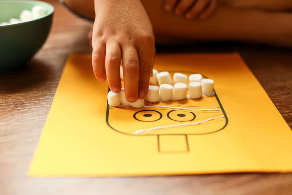 Mini marshmallows being glued to a pumpkin craft by a child's hand.