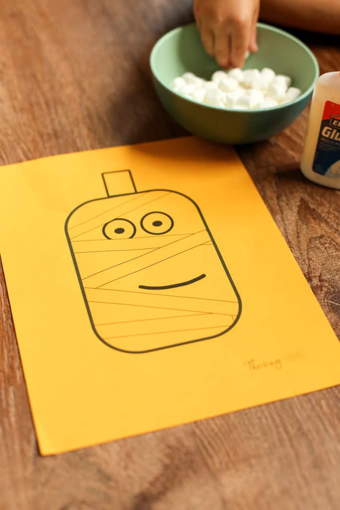 Mummy pumpkin printable template on orange paper with a bowl of mini marshmallows and school glue sitting beside it.