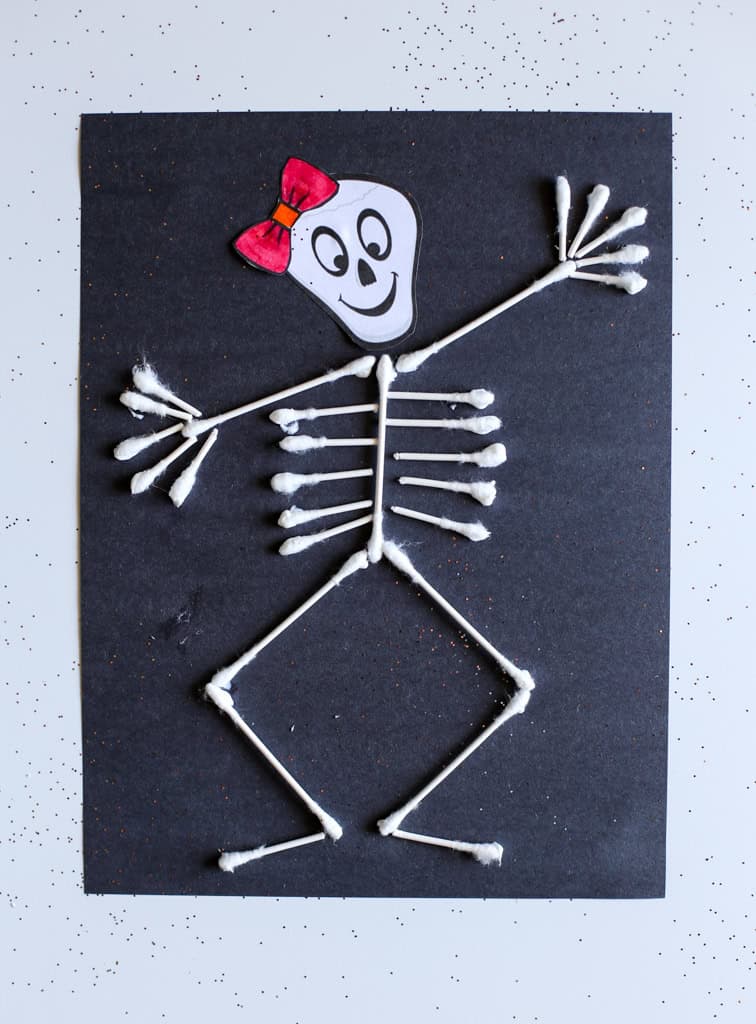 Q-tip skeleton on black construction paper with a cutout skull with a red bow.