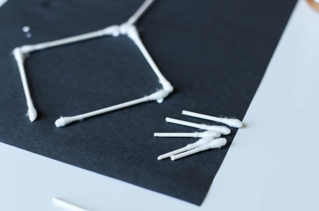 Q-tips as legs of a skeleton on a black piece of paper with some unused Q-tips already snapped in half.