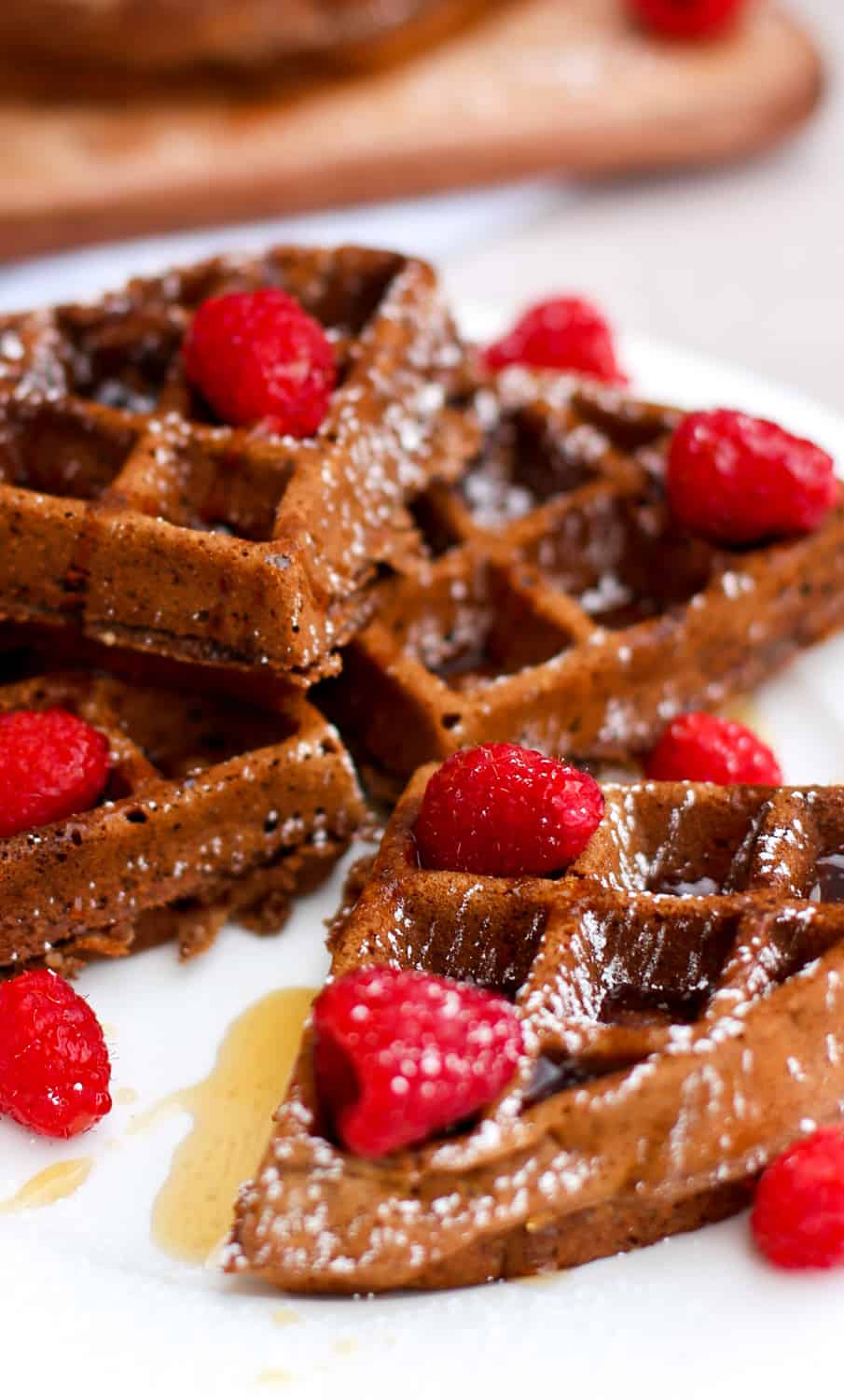Healthy chocolate waffles stacked on a plate topped with raspberries and powdered sugar.
