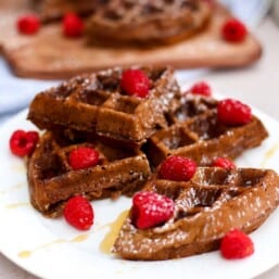 Waffles stacked on a plate with raspberries on top and drizzled with syrup.