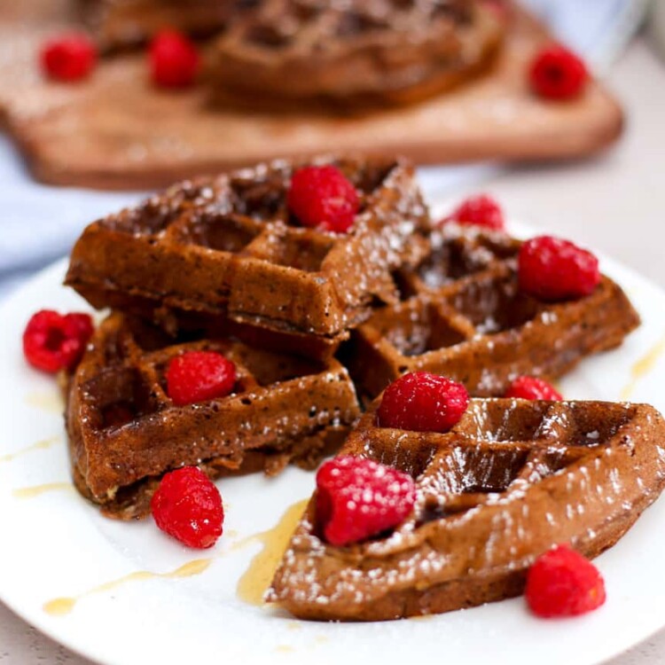 Waffles stacked on a plate with raspberries on top and drizzled with syrup.