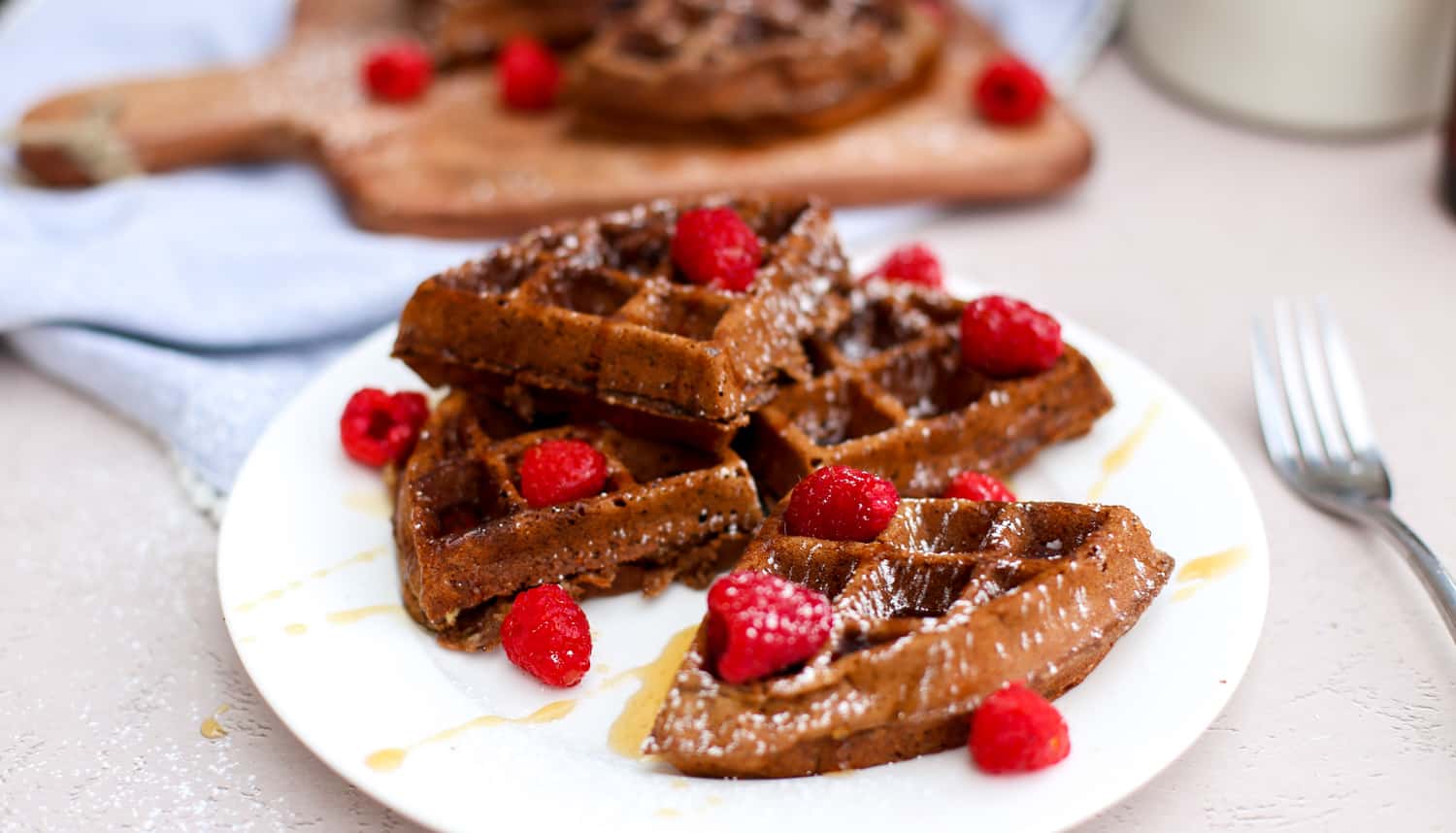 Chocolate waffles broken up and stacked on a white plate with raspberries on top and drizzled with syrup.