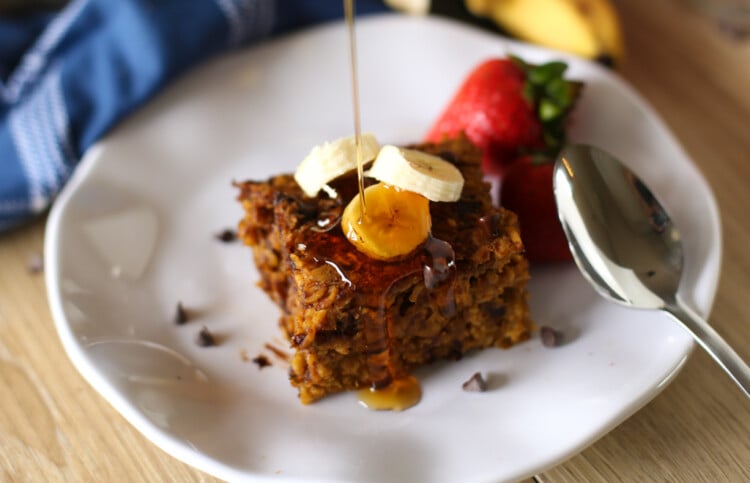 A serving of pumpkin baked oatmeal on a plate with a few banana slices and syrup being poured on top.