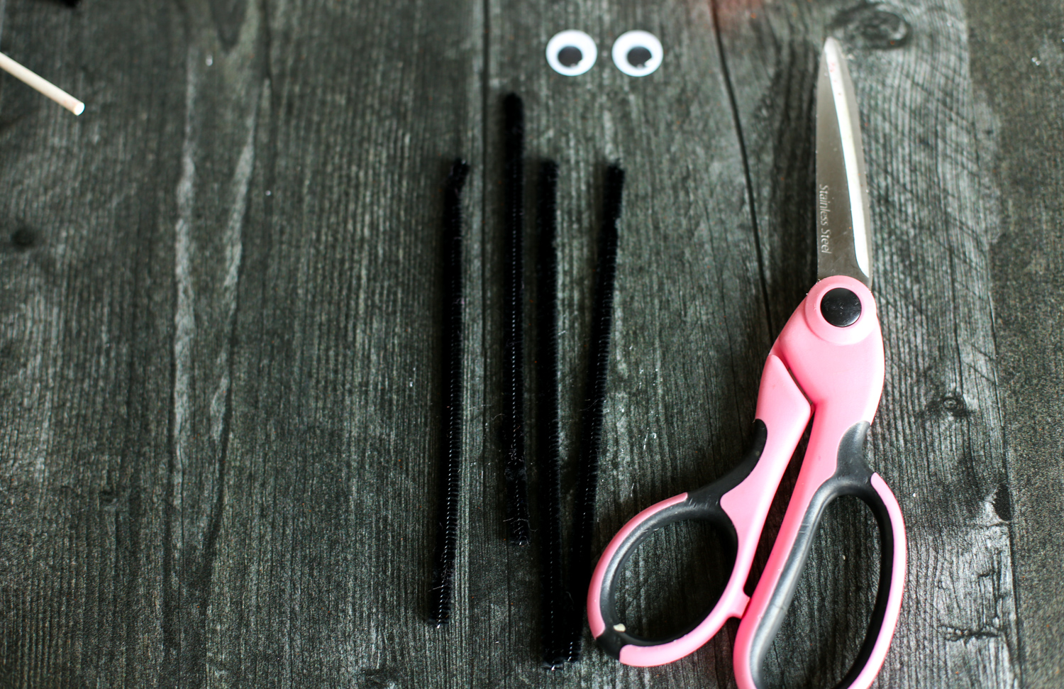 Black pipe cleaners cut in half, googly eyes, and a pair of scissors laid out on a wooden table.