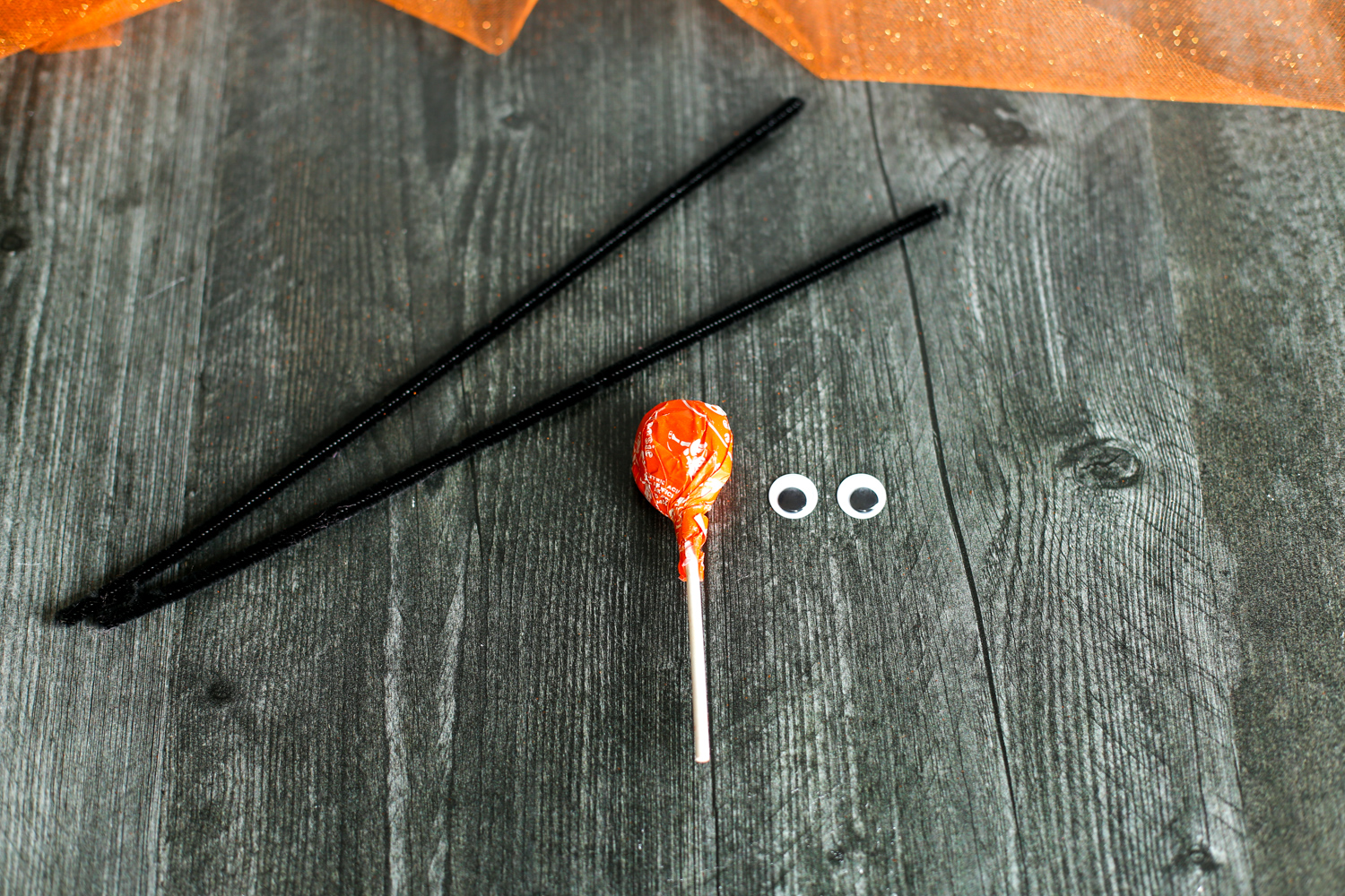 Pipe cleaners, tootsie roll pop and googly eyes laid out on a wooden table.