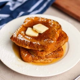 Pumpkin french toast stacked on a plate with two tabs of butter on top.