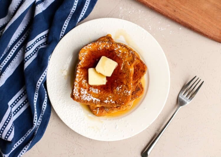 Two slices of pumpkin french toast stacked in a white plate with maple syrup and pats of butter on top.