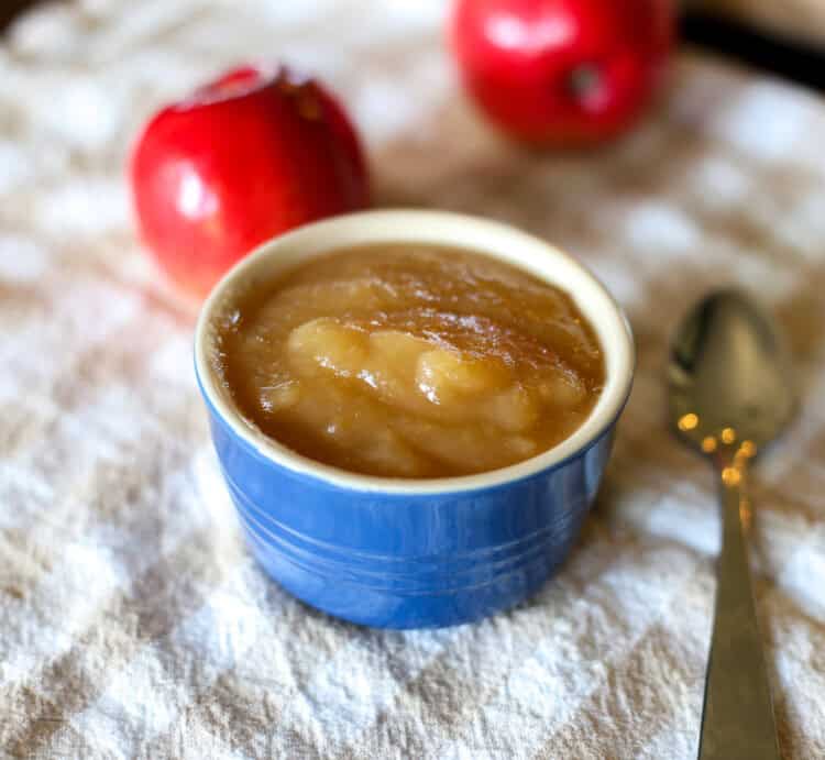 Slow cooker applesauce in a small blue bowl with a spoon next to it and apples in the background.