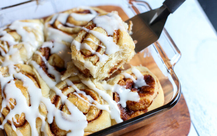 Baked cinnamon rolls with icing drizzled and a spatula lifting the first one out of the dish.