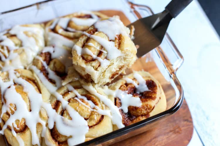 Baked cinnamon rolls with icing drizzled and a spatula lifting the first one out of the dish.