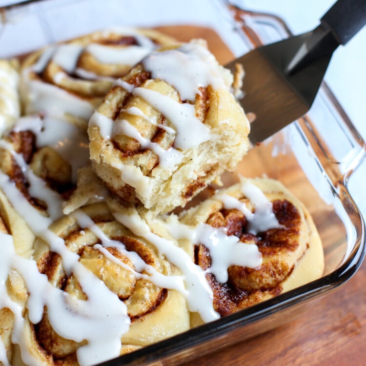 Baked cinnamon rolls with icing drizzled and a spatula lifting the first one out of the dish.