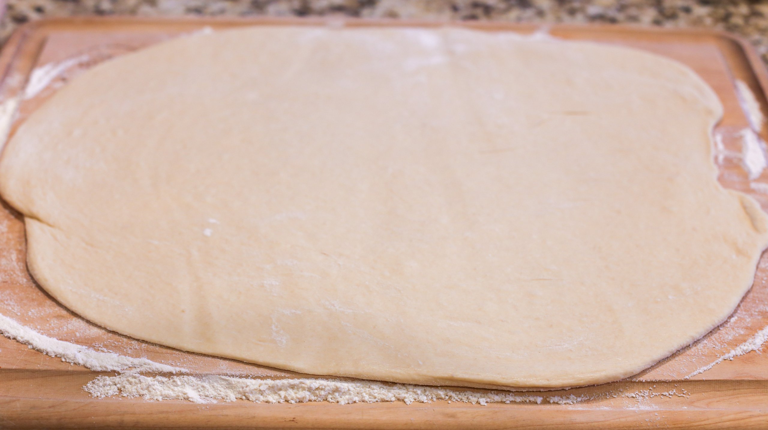 Cinnamon roll dough rolled flat in an oval on wooden cutting board.