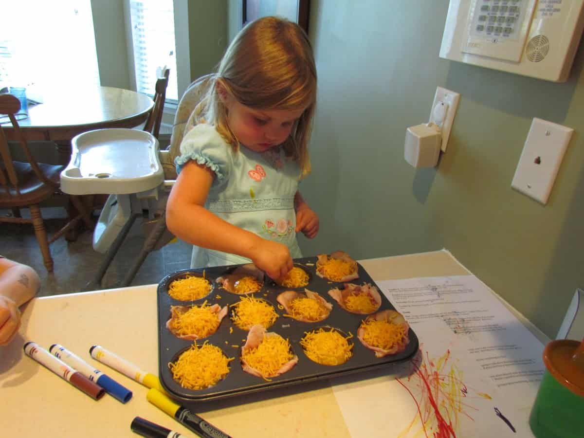 Baked Egg Cups Young child putting shredded cheese on top of ham and cheese egg muffins.