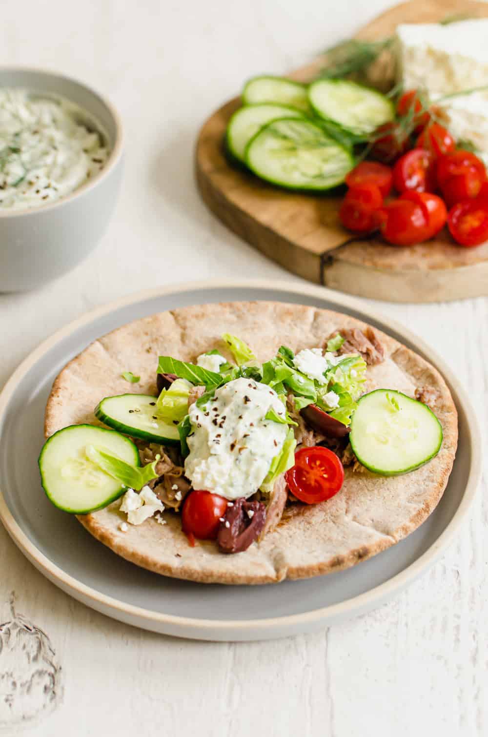 A pulled pork pita wrap on a blue plate with a bowl of tzatziki sauce and board of vegetables in the background.