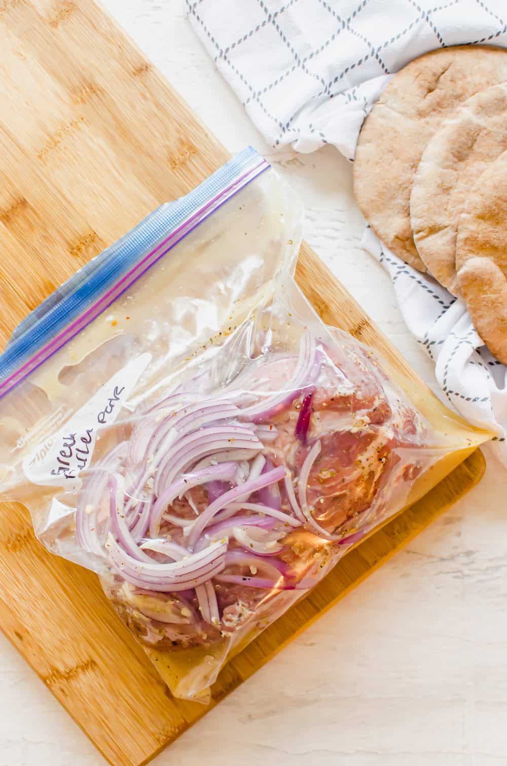 A freezer bag labeled Greek pulled pork with a raw pork shoulder, red onion slices, chicken broth, and Greek seasonings.