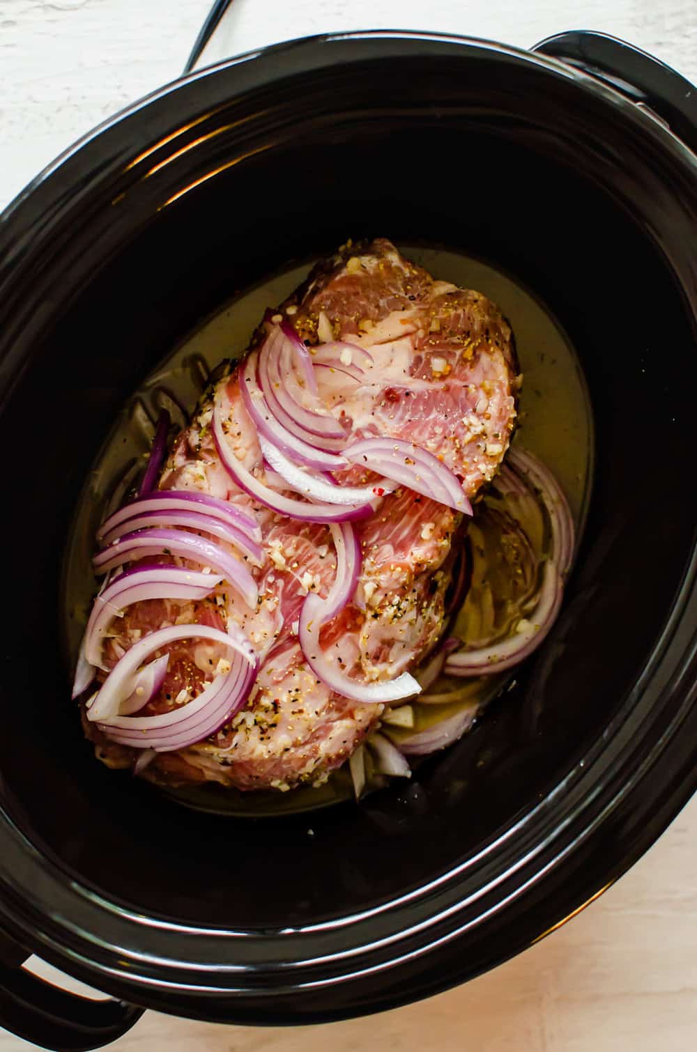 Raw pork shoulder in a slow cooker with Greek seasonings, chicken broth, and red onion slices.