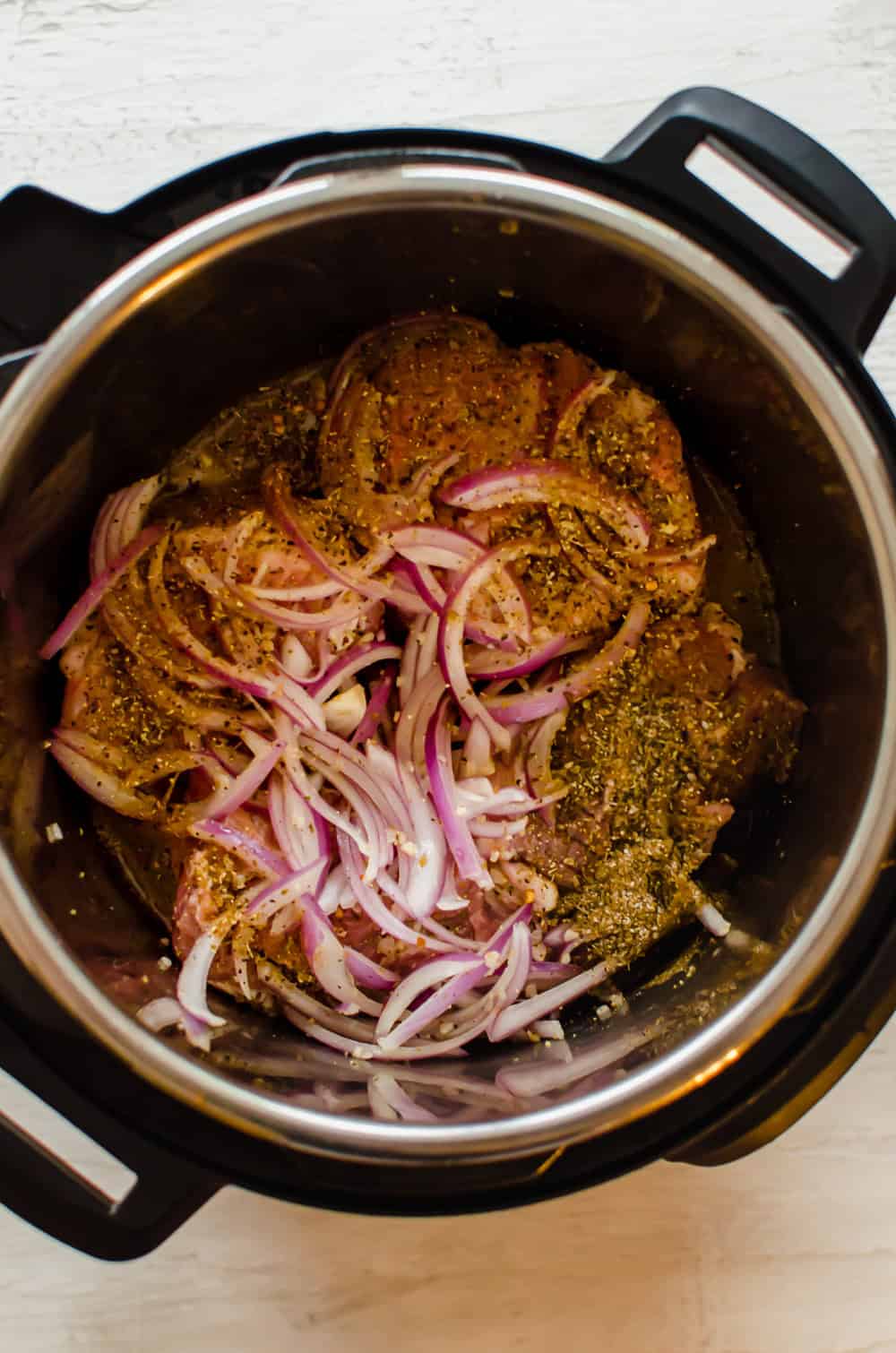 A pork shoulder cut in 4 equal chunks in an Instant Pot with Greek seasonings, red onion slices, and chicken broth.