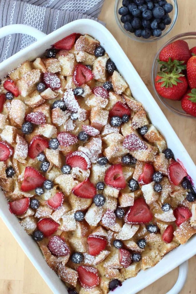 Berry french toast casserole in a casserole dish with sliced strawberries, blueberries, and powdered sugar on top.