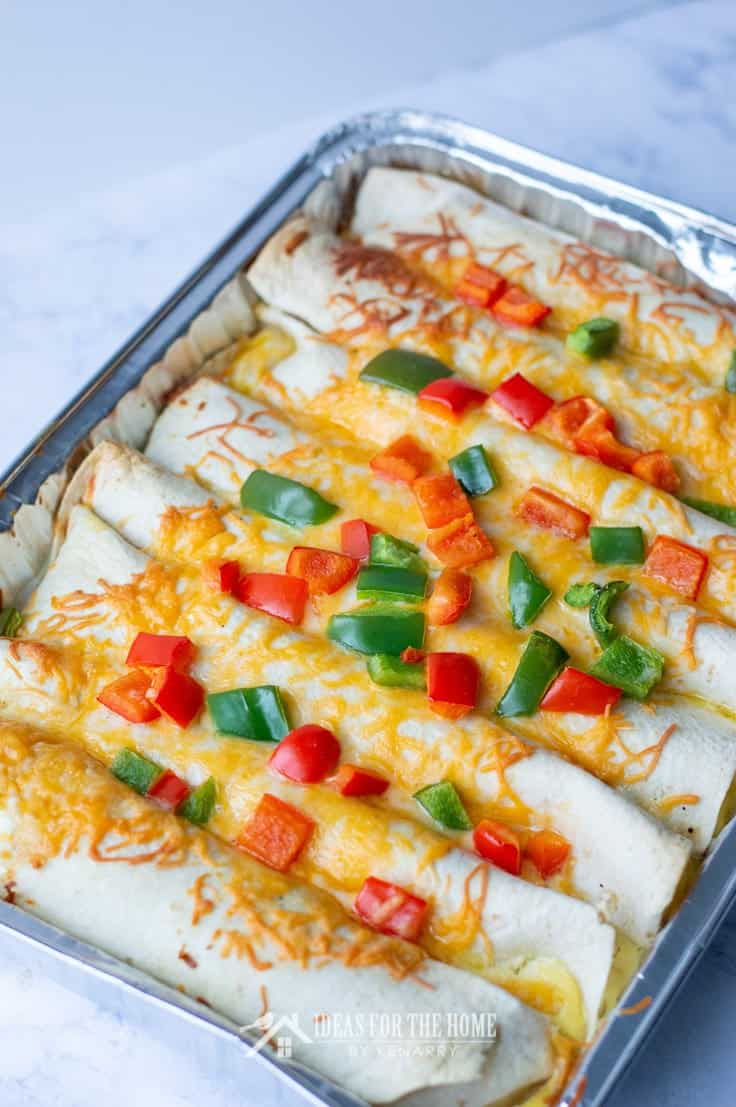 Breakfast enchiladas lined up in a baking pan with chopped green and red peppers on top.