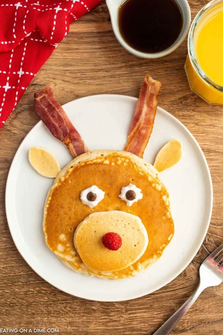 Different size pancakes made into a reindeer face with bacon as antlers and a raspberry as its nose.