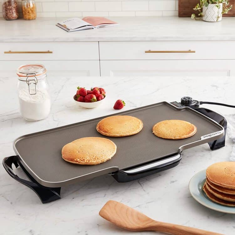 An electric griddle on a counter with three pancakes cooking on it, and a stack of pancakes on a plate next to it.