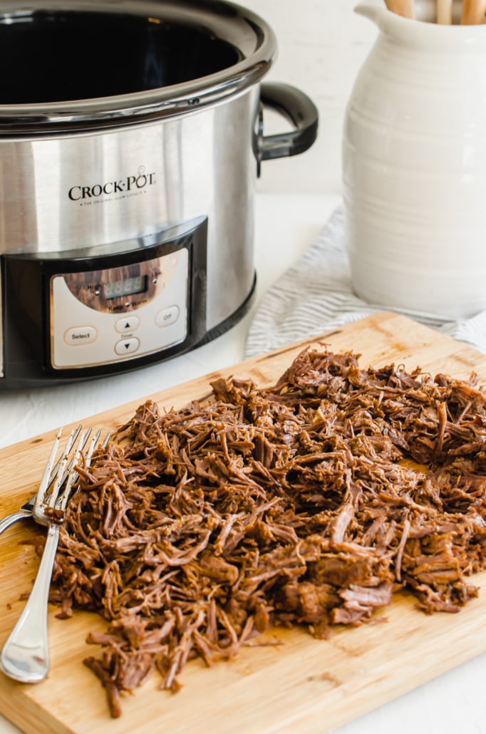 shredded beef on a wooden cutting board with a crockpot in the background