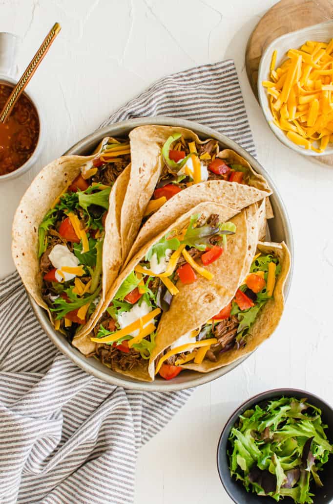 Shredded beef tacos in a metal pan with bowls of lettuce and cheese on the side. 