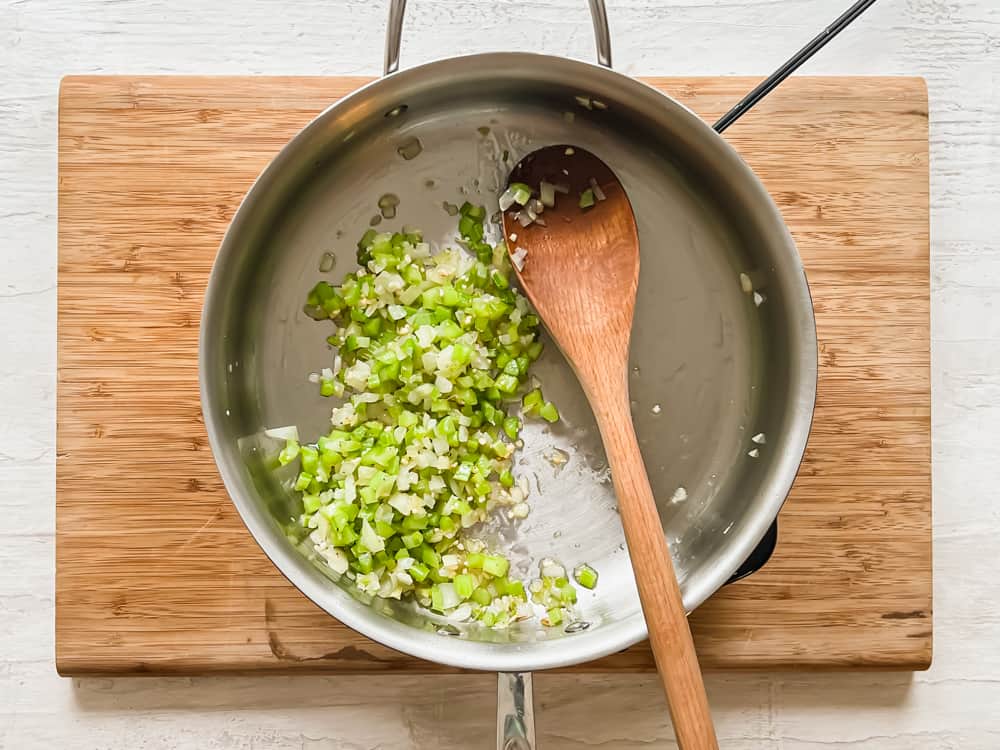 Celery and onion sauteing in a stainless steel pan with a wooden spoon stirring.