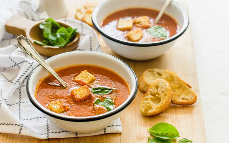 Two bowls of tomato soup with croutons and fresh basil on top.