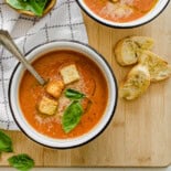Bowl of tomato bisque soup with crusty bread, fresh basil leaves, and Parmesan on top.