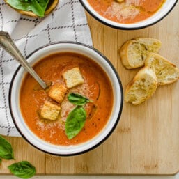Bowl of tomato bisque soup with crusty bread, fresh basil leaves, and Parmesan on top.