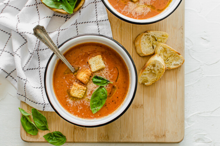 Bowl of tomato bisque soup with crusty bread, fresh basil leaves, and Parmesan on top.