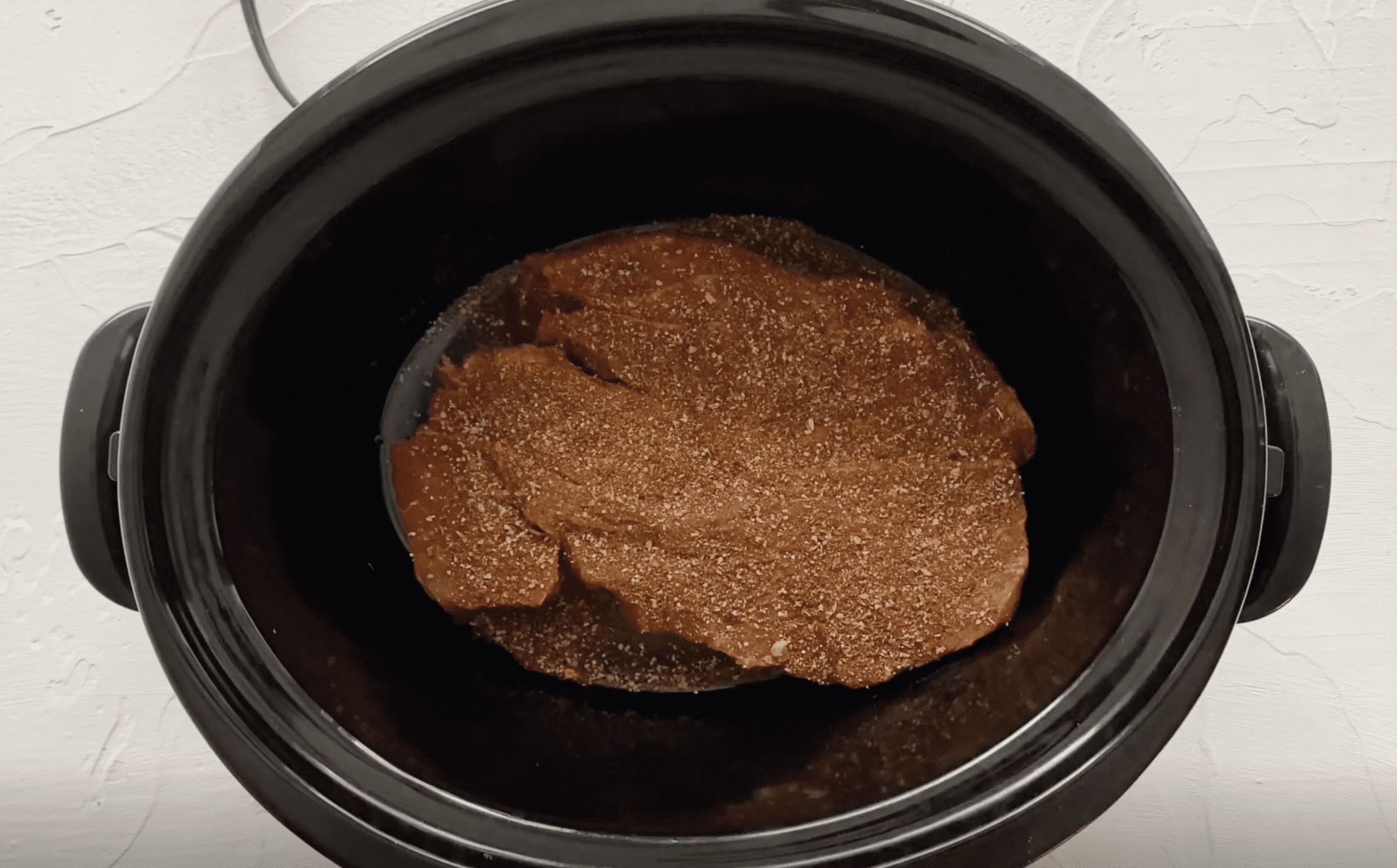An uncooked chuck roast with seasonings on it in a slow cooker.