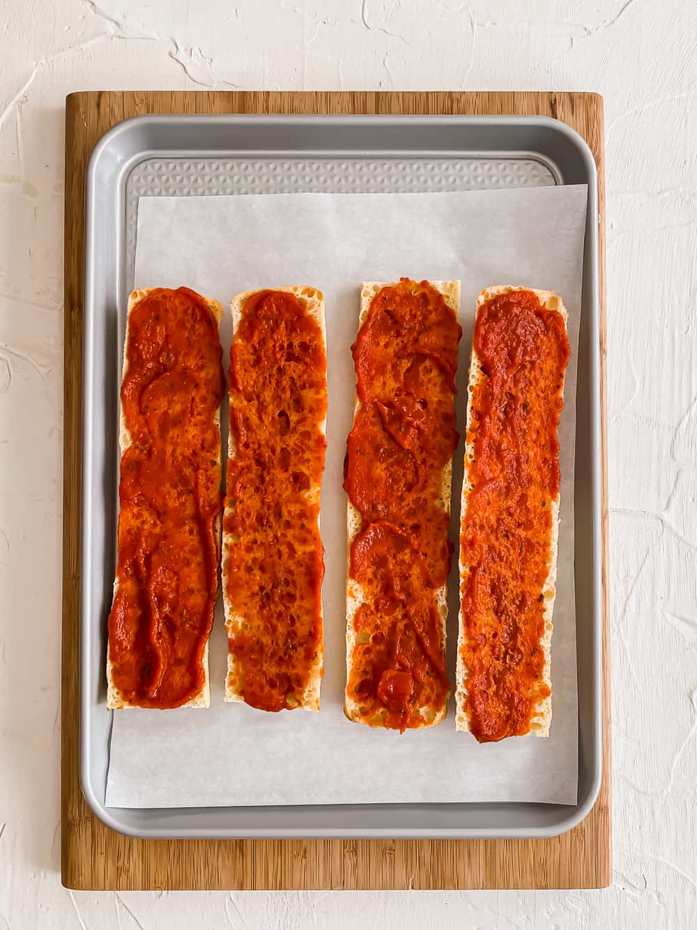 Sliced baguettes on a baking sheet with pizza sauce on them.