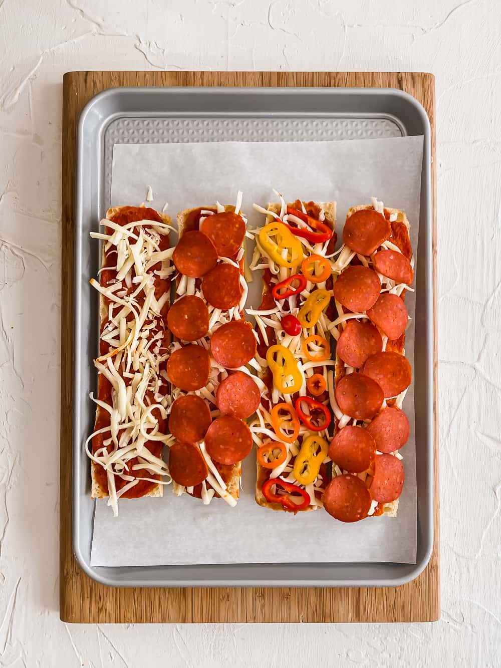 Sliced baguettes on a baking sheet with pizza sauce, shredded mozzarella, and different toppings on each.