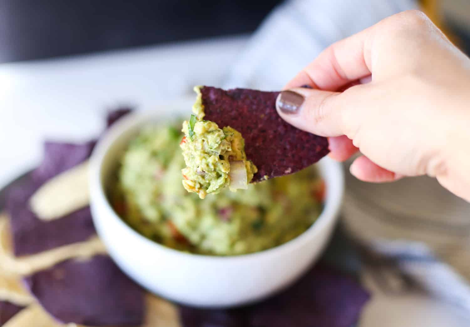 A hand dipping a blue corn chip into a bowl of guacamole.